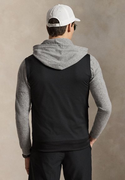 Άνδρας με μαύρο αμάνικο γιλέκο πάνω από γκρι φούτερ και λευκό καπέλο μπέιζμπολ, στέκεται προς ουδέτερο μπεζ φόντο.