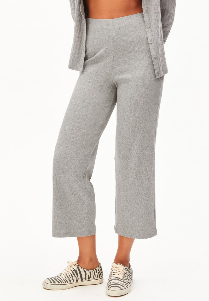 Person trägt eine hoch taillierte, graue Hose mit weitem Bein und zebragestreifte Sneakers, dazu ein teilweise sichtbarer grauer Cardigan, steht vor weißem Hintergrund.