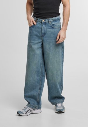 Jeans en denim larges de couleur bleu clair avec un bouton frontal unique, des passants de ceinture et des poches latérales, associés à des chaussures de sport grises et blanches.
