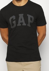 Una camiseta de algodón negra presenta un gran logo en gris oscuro de "GAP" en el pecho. El diseño incluye un cuello redondo clásico y mangas cortas.