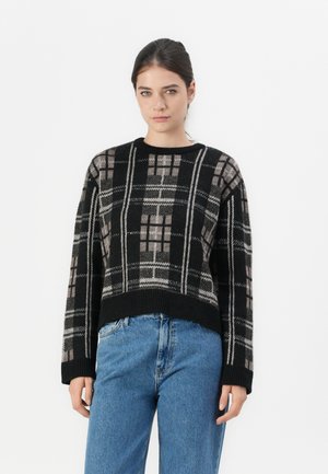 Črno-siva pletena jopica z geometrijskim vzorcem in okroglim izrezom ter rebrastimi robovi, v kombinaciji z modrimi jeans hlačami z visokim pasom.