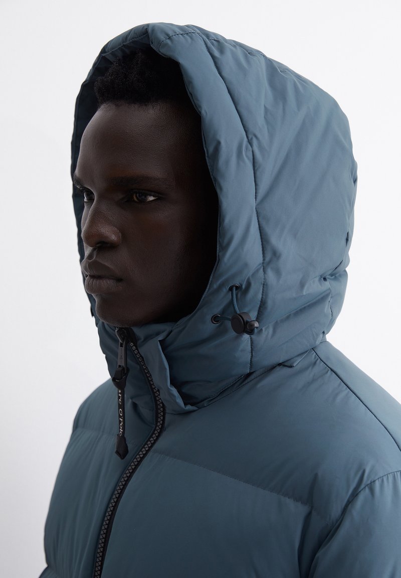 Daunenjacke mit dunkelblauem Außenmaterial, ausgestattet mit einem hohen Kragen und einer verstellbaren Kapuze mit Kordelzug. Glatte Textur mit durchgehendem Reißverschluss an der Vorderseite.