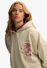 Beżowy hoodie z kapturem, zdobiony haftem w postaci czerwonego i złotego smoka z przodu. Miękka tkanina oraz luźny krój.