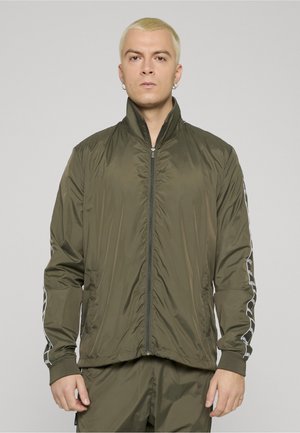 Mann med kort blondt hår iført olivengrønn treningsjakke med glidelås og matchende bukser med mønstrede striper på ermene, står foran en ensfarget bakgrunn.