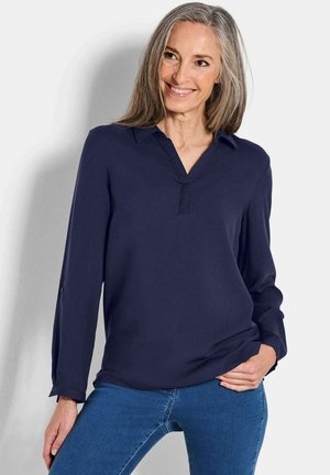 Marineblauwe, lange mouw blouse met een V-hals en een voorste knoopsluiting. Zacht materiaal met een lichte glans en met knoopjes aan de manchetten. Gecombineerd met blauwe jeans.