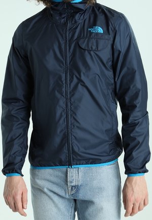 Ciemnoniebieska, lekka kurtka z zamkiem z przodu, elastycznymi mankietami i kieszenią na piersi, z niebieskim logo po lewej stronie.