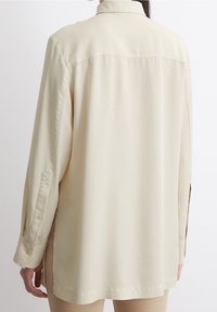 Camicia beige a maniche lunghe con colletto e polsini abbottonati, caratterizzata da una linea dritta e spacchi laterali per facilitare i movimenti. Texture del tessuto liscia.