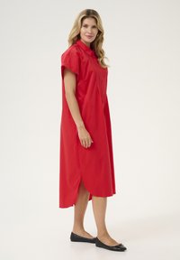 Abito di cotone rosso con colletto, maniche corte, chiusura con bottoni e spacchi laterali. Scarpe nere basse completano l'outfit.