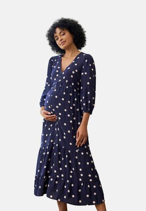 Zwangere vrouw met krullend haar, gekleed in een marineblauwe midi-jurk met witte stippen, staand en zachtjes haar buik vasthoudend.