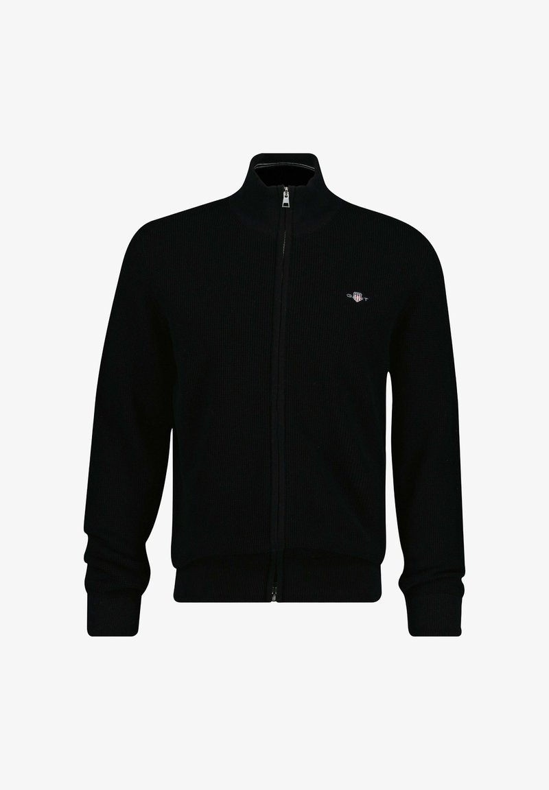 Chaqueta negra de punto con cierre de cremallera, cuello alto, textura acanalada y detalle de logo en el pecho izquierdo. Cuenta con mangas largas y diseño ajustado.
