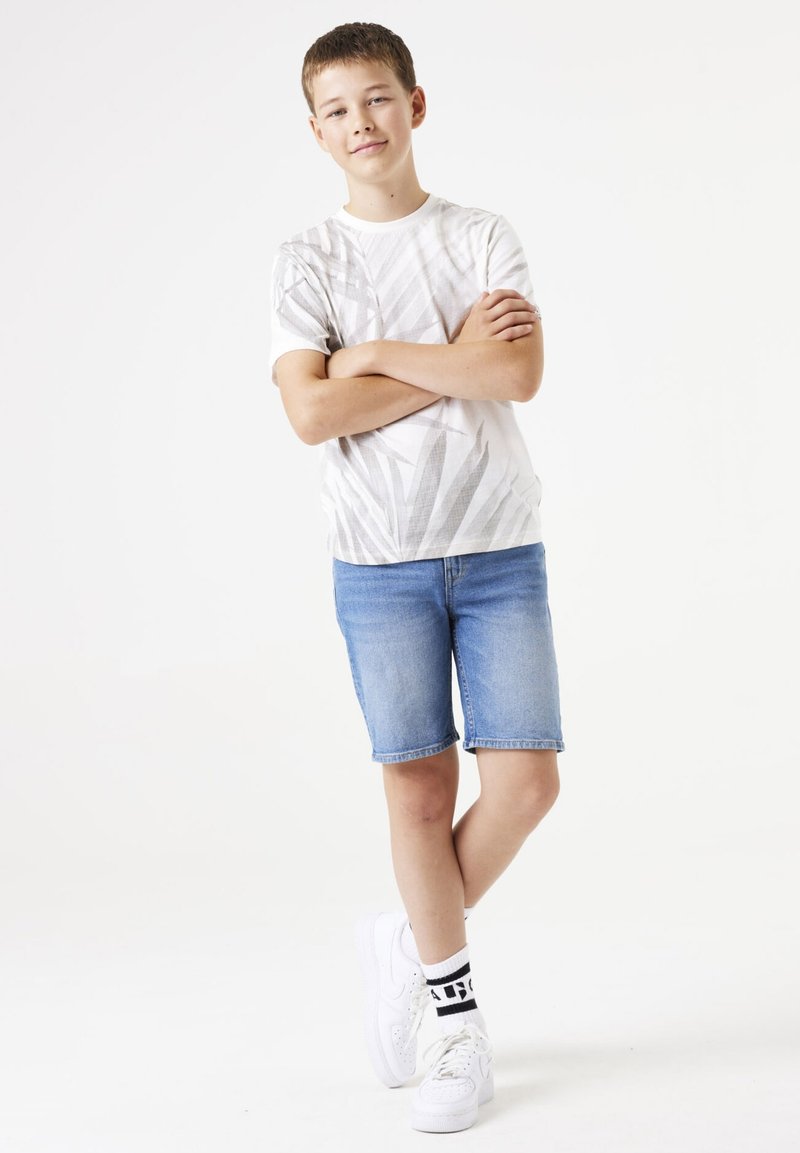 Witte korte mouwen t-shirt met grijs bladpatroon, gecombineerd met lichtblauwe denimshorts en witte sneakers met zwarte accenten.