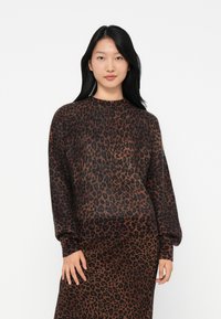 Jovem mulher com cabelo preto longo, vestindo um suéter e uma saia de estampa de leopardo em tons castanho escuro e preto, de pé contra um fundo branco.
