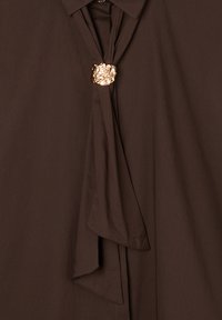 Blusa marrone scuro con un dettaglio drappeggiato sul davanti, fissata da una chiusura decorativa in tono oro texture nel collo.