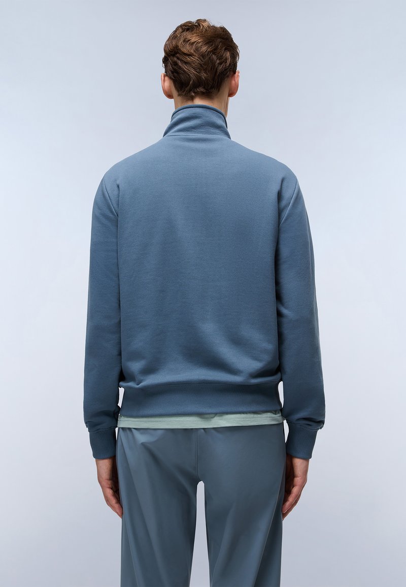 Sudadera azul de peso medio con cuello alto y puños acanalados, con una textura suave y un corte relajado, usada sobre capas de color verde claro.