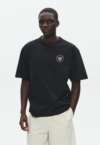 Νεαρός άνδρας που φοράει μαύρο T-shirt με λευκό κυκλικό λογότυπο στο αριστερό στήθος και ανοιχτόχρωμο παντελόνι, στέκεται μπροστά σε ένα απλό λευκό φόντο.