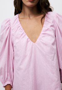 Hellrosa Bluse mit tiefem V-Ausschnitt, ausgestattet mit Puffärmeln und gerafften Details an den Schultern und Bündchen. Glatte Stofftextur.