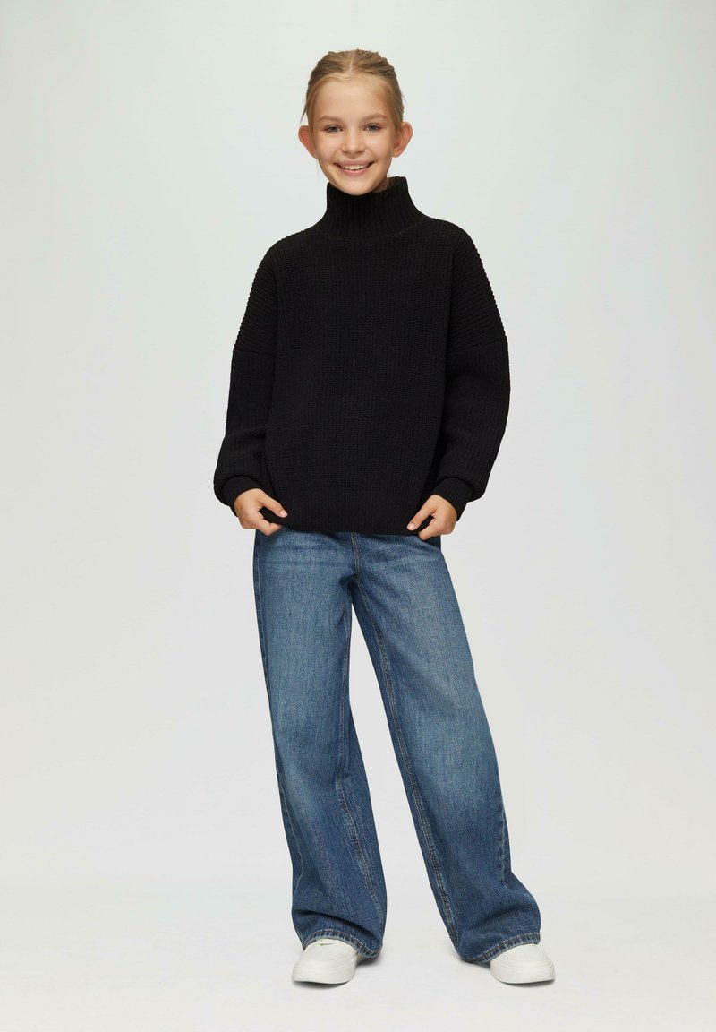 Zwarte gebreide coltrui met ribbelstructuur, gecombineerd met ruime blauwe jeans en witte sneakers. Eenvoudige en casual outfit.