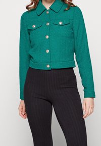Tangerine-farbene, kurzwaarige Jacke mit strukturiertem Finish, die über einen Kragen, zwei Brusttaschen und Knopfverschlüsse in Goldton verfügt. Kombiniert mit schwarzen, gerippten Hosen.