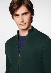 Ciemnozielony, rozpinany sweter dzianinowy z teksturowanym wzorem, wysoki kołnierz i srebrny zamek, nałożony na granatową koszulę.