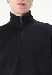 Polo noir à fermeture éclair avec un col montant, une texture côtelée et une fermeture éclair en métal à l'avant. Design simple et ajusté. Aucun motif visible.