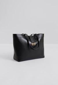 Μαύρη δερμάτινη τσάντα tote με ευρύ άνοιγμα στο πάνω μέρος, λεπτές λαβές και χρυσό διακοσμητικό μπροστά. Ομαλή υφή και δομημένο σχήμα.