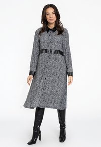 Schwarz-weiß gestreiftes Kleid im Chevron-Muster mit durchgehender Knopfleiste, Hemdkragen, schwarzem Lederbund und langen Ärmeln. Getragen mit kniehohen schwarzen Stiefeln.