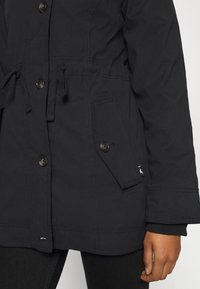 Veste noire avec fermeture à boutons, cordons ajustables à la taille, poche à rabat avec bouton, portée sur un pantalon noir, main reposant sur le côté.