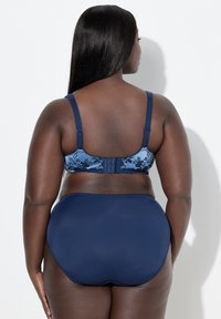 Mujer de piel oscura y cabello largo y liso lleva un sujetador floral azul marino y braguitas de cintura alta a juego, de frente a una pared blanca.