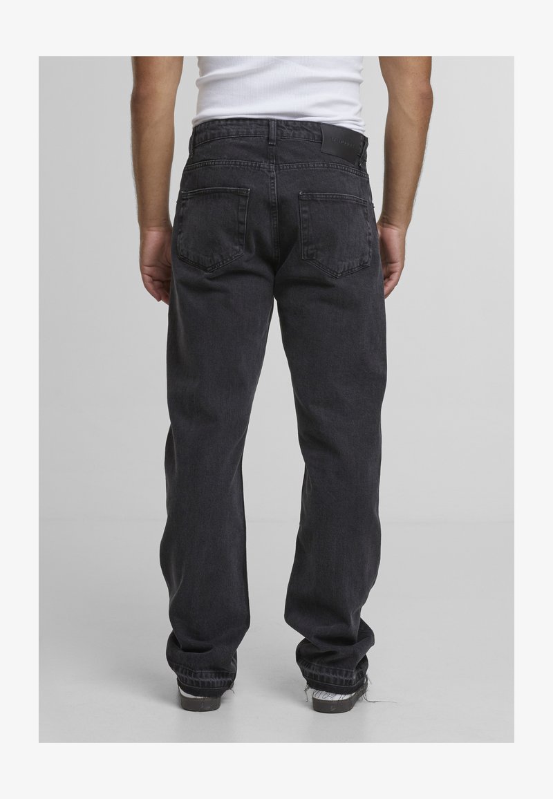 Mann in locker sitzender schwarzer Jeans und weißen Turnschuhen, von hinten vor einem einfarbigen grauen Hintergrund gezeigt.