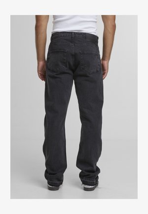 Mand iført løstsiddende sorte jeans og hvide sneakers, vist bagfra mod en ensfarvet grå baggrund.