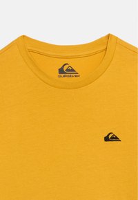 Μουσταρδί κίτρινο t-shirt με στρογγυλή λαιμόκοψη, μικρό μαύρο λογότυπο Quiksilver στο στήθος και τυπωμένη επωνυμία της μάρκας στο εσωτερικό του λαιμού.