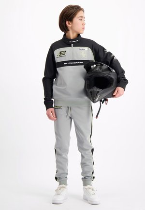 Grijs en zwart trainingspak met een rits-jack en bijpassende broek, voorzien van groene accenten en logodetails, gecombineerd met een zwarte helm.