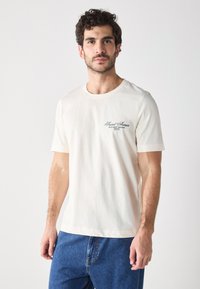 Cream cotton t-shirt with short sleeves, featuring a rounded neckline and a small green logo on the left chest area.