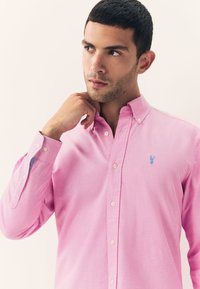 Light pink button-up shirt made of cotton, featuring a blue embroidered logo on the chest and contrasting inner cuff detail.