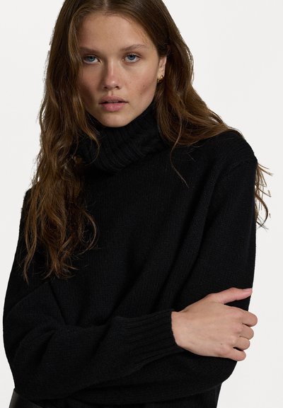 Pulover negru din tricot cu guler tip tortiță. Mâneci lungi, croială lejeră și țesătură texturată. Design simplu, fără modele vizibile sau accesorii.