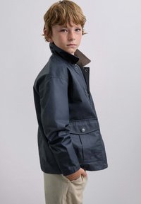 Niño pequeño con cabello rojo y pecas, vestido con una chaqueta azul oscura con collar alto y pantalones beige, de pie con una mano en el bolsillo.