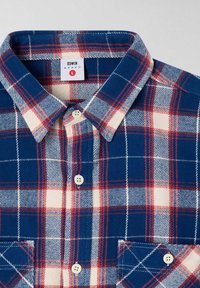 Camicia di flanella a quadri blu e bianchi con accenti rossi, caratterizzata da un colletto classico, chiusura frontale con bottoni e due tasche sul petto. Etichetta del marchio visibile.