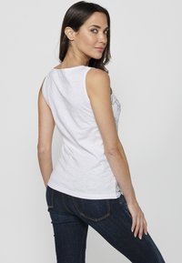 Top bianco senza maniche con un design aderente, caratterizzato da un motivo grafico evidenziato sul lato, abbinato a jeans in denim blu scuro.