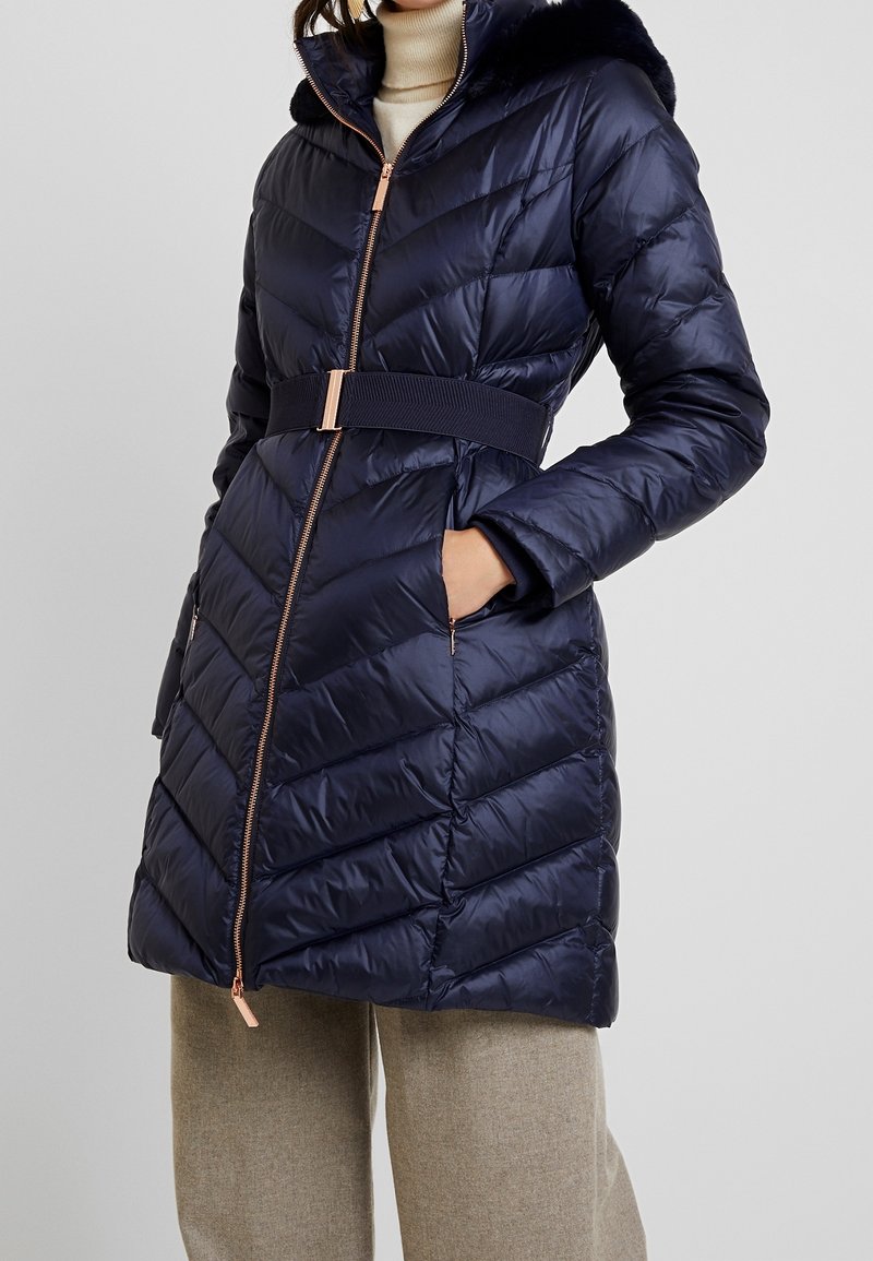 Abrigo puffer navy con capucha forrada de piel sintética, cintura ceñida y patrón de rombos. Cuenta con un cierre completo y bolsillos laterales.