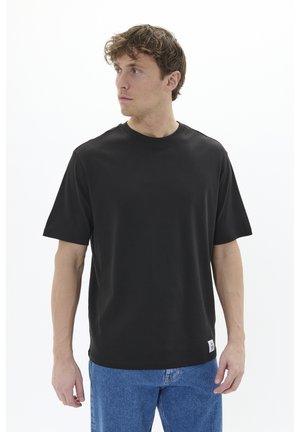 Schwarzes T-Shirt mit kurzen Ärmeln aus weichem Stoff, Rundhalsausschnitt und einem kleinen Etikett an der unteren linken Seite. Kombiniert mit blauen Jeans.