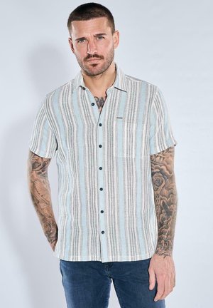 Mann mit kurzen Haaren und Tattoos, der ein kurzärmliges Hemd mit vertikalen Streifen und dunkelblauen Jeans trägt, steht vor einem einfarbigen Hintergrund.