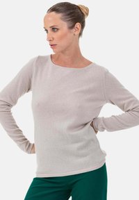 Femme aux cheveux relevés en chignon, portant un pull beige clair à manches longues et un pantalon vert foncé, posant les mains sur les hanches et regardant vers la gauche.