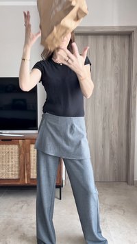 Pantaloni grigi su misura con una vestibilità ampia e sovrapposizione asimmetrica a strati, abbinati a una maglietta nera attillata a maniche corte. Busta di carta marrone in mano.