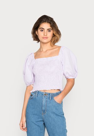 Top curto de padrão xadrez roxo com mangas bufantes e decote quadrado. Textura suave, cintura franzida e barra desfiada. Combinado com jeans azuis.