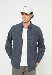 Fiatal férfi fehér sapkával, amelyen a "BOSS" felirat olvasható, tengerészkék kockás gombos ingben, fehér póló fölött, és szürke nadrágban, egyszínű háttér előtt.