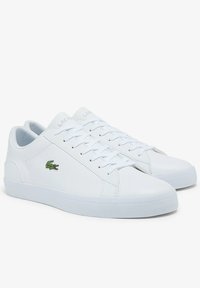 Baskets blanches en matériau synthétique, avec une finition lisse, des lacets blancs plats et un logo de crocodile brodé vert sur le côté.
