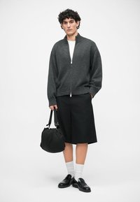 Σκούρο γκρι φούτερ με φερμουάρ πάνω από λευκό πουκάμισο, σε συνδυασμό με μαύρες σορτς μέχρι το γόνατο, λευκές κάλτσες, μαύρα παπούτσια και μαύρη τσάντα. Απλός σχεδιασμός.