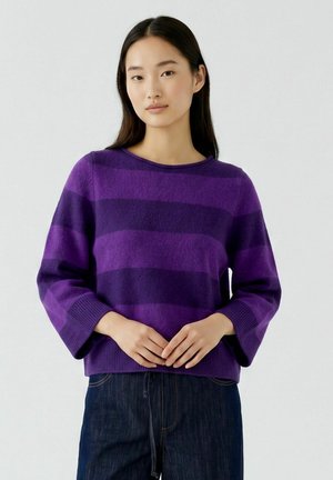 Jeune femme aux longs cheveux foncés portant un pull rayé violet et un jean foncé, debout les mains jointes devant un fond uni.