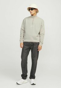 Light gray zip-up sweatshirt, black jeans, white sneakers, and a white bucket hat. Model wears black sunglasses. Simple design, casual fit.