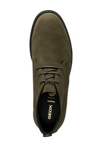 Zapato de ante verde oliva con puntera redonda, diseño de cordones y suela de goma negra. Presenta un logotipo dorado en la plantilla y una textura superior lisa.